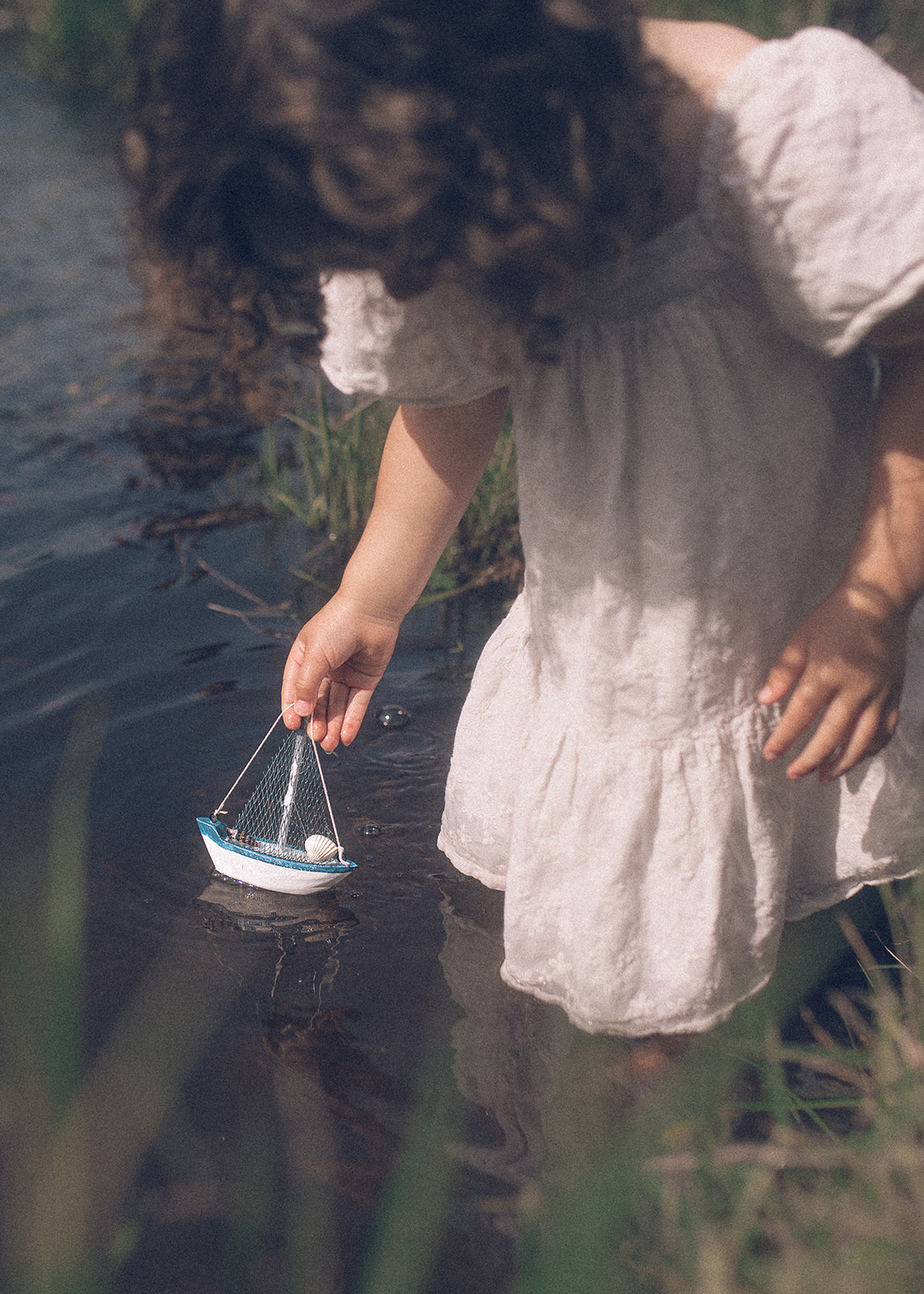 A toddler girl in a white dress places a tiny boat in a slow creek after visiting this toy store in Hamilton, MT