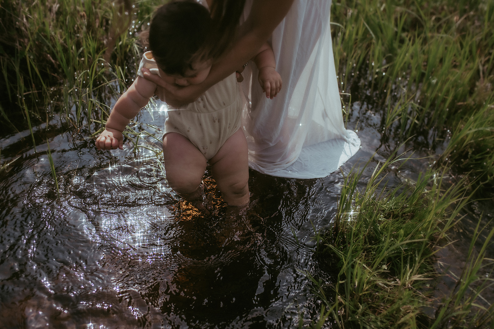 An infant walks in a shallow creek among the grass with mom's help
