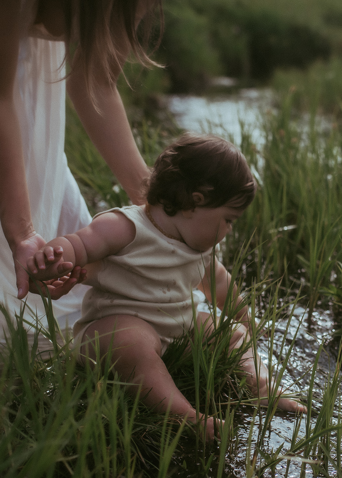 A mother helps her infant sit on the edge of a creek while reaching for the water in a white onesie after visiting Happy Kid Store