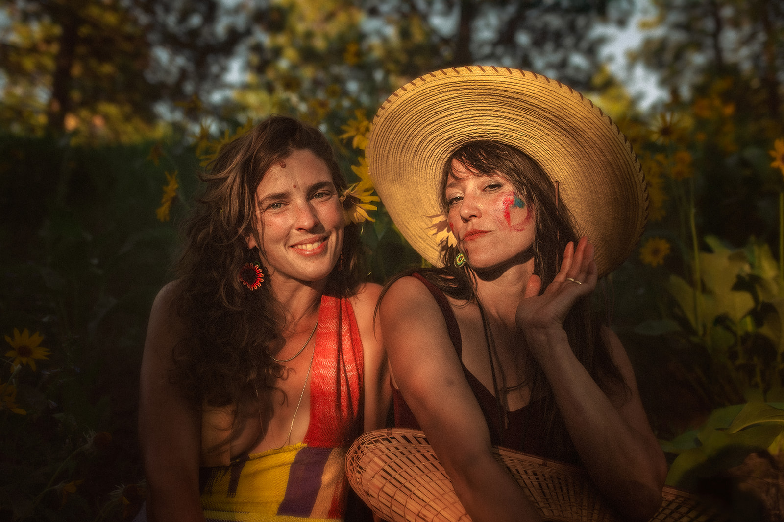 Two women with face paint and colorful dresses and big hats sit among wildflowers in a forest after finding OBGYN in Hamilton, MT