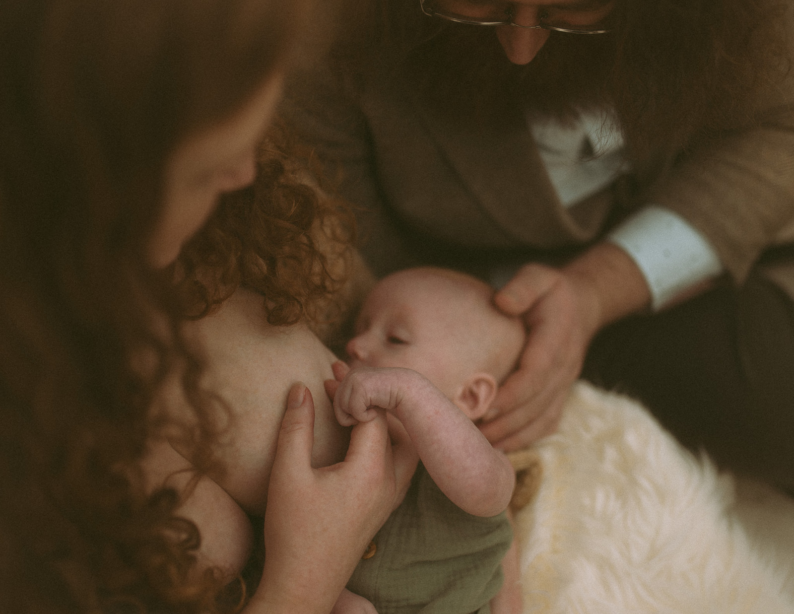 New parents breastfeed their infant while sitting together with dad's hand on baby's head