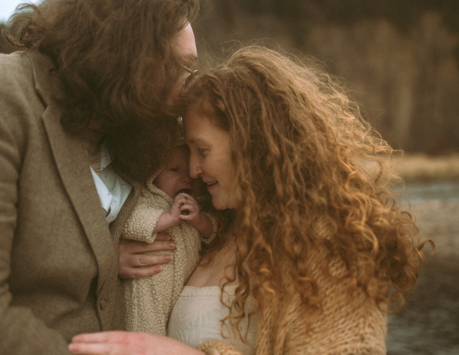 Happy new parents snuggle their infant by a river in a knit sweater and brown sports coat