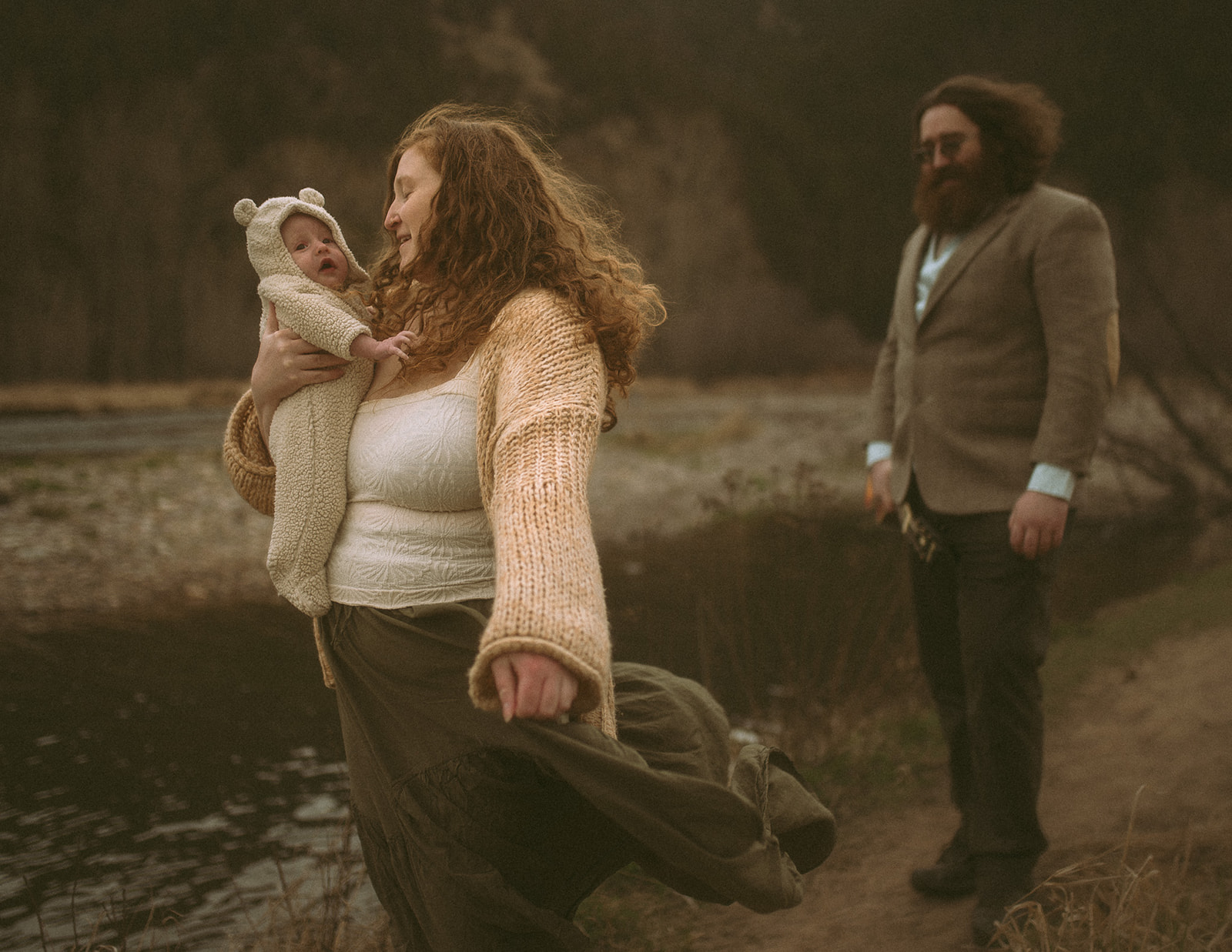 A mom and dad explore a river's edge with their infant in mom's arms on a windy day after visiting pediatrician in Hamilton, MT