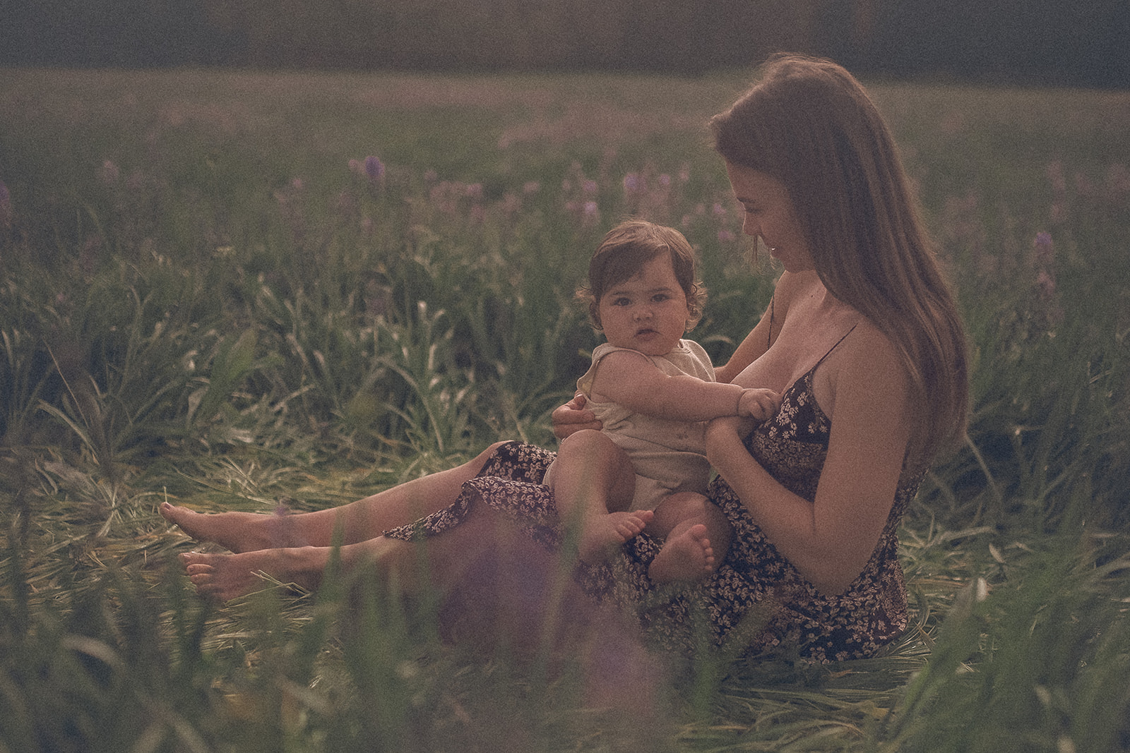 A mother in a black floral dress sits in a meadow with her infant in her lap after visiting Yours Mine And Ours Kid Store