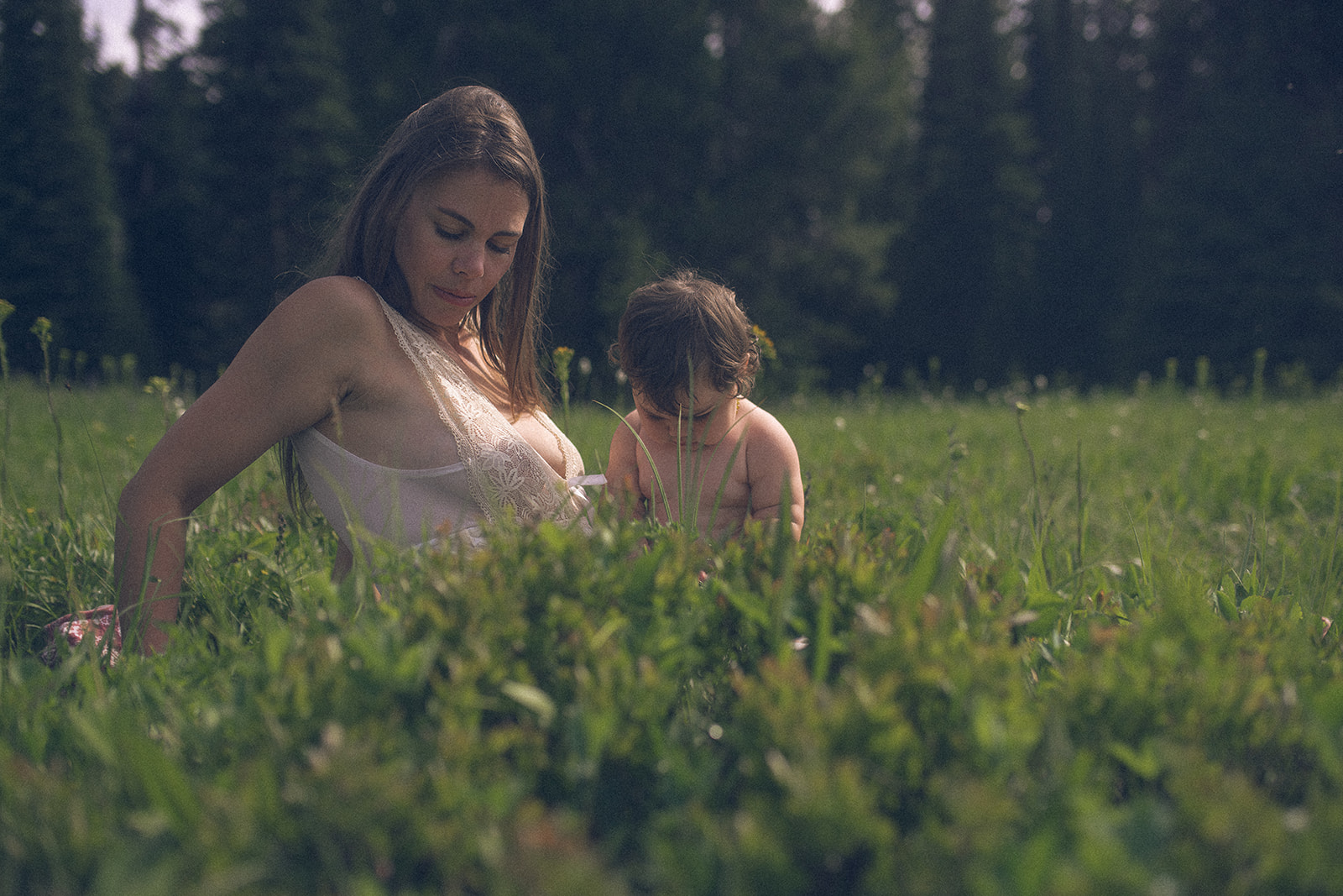 A mom in a white dress sits with her baby in a meadow