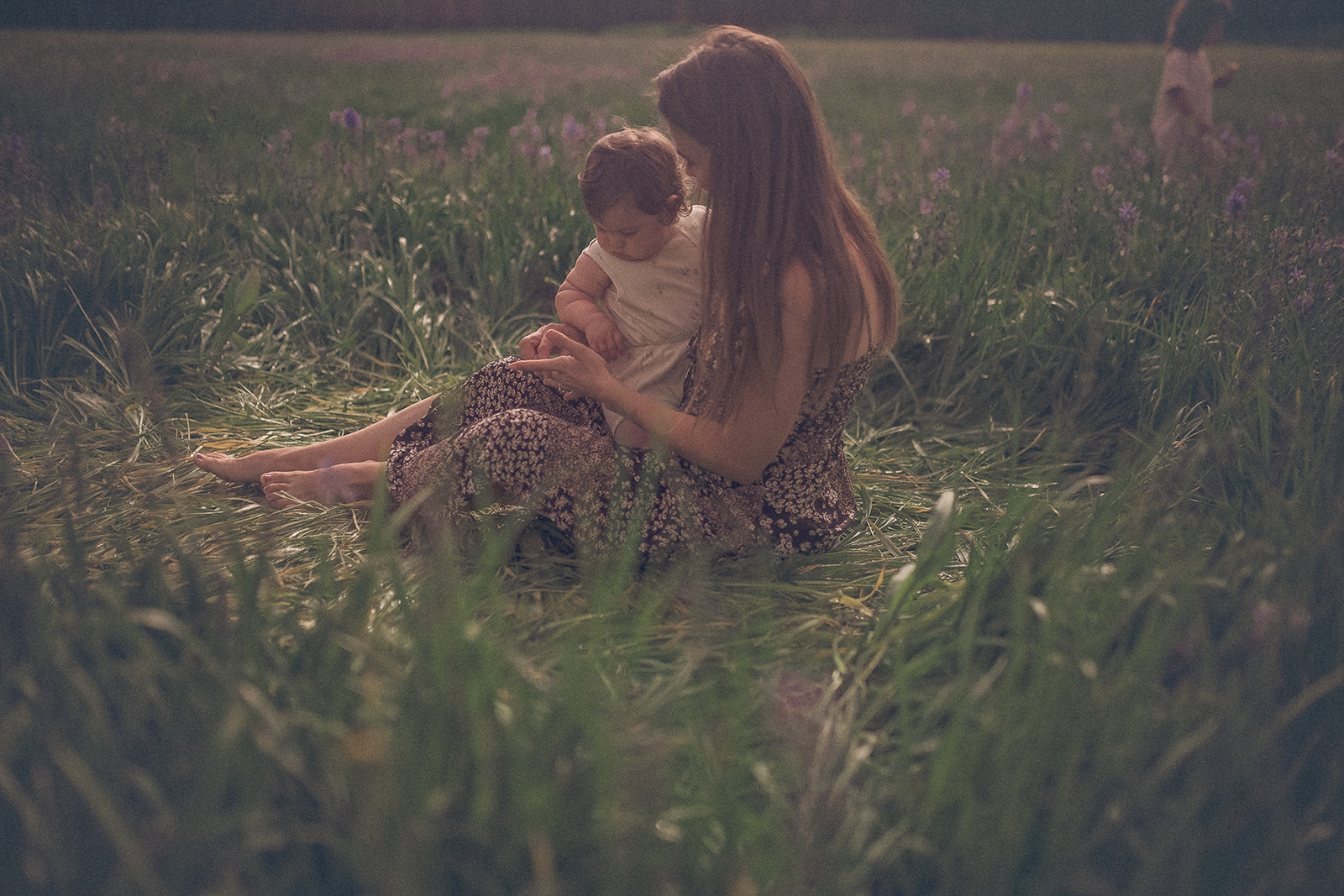 A happy mom sits in a patch of laid down grass exploring wildflowers with her infant in a white dress in her lap after some baby music classes in Hamilton, MT