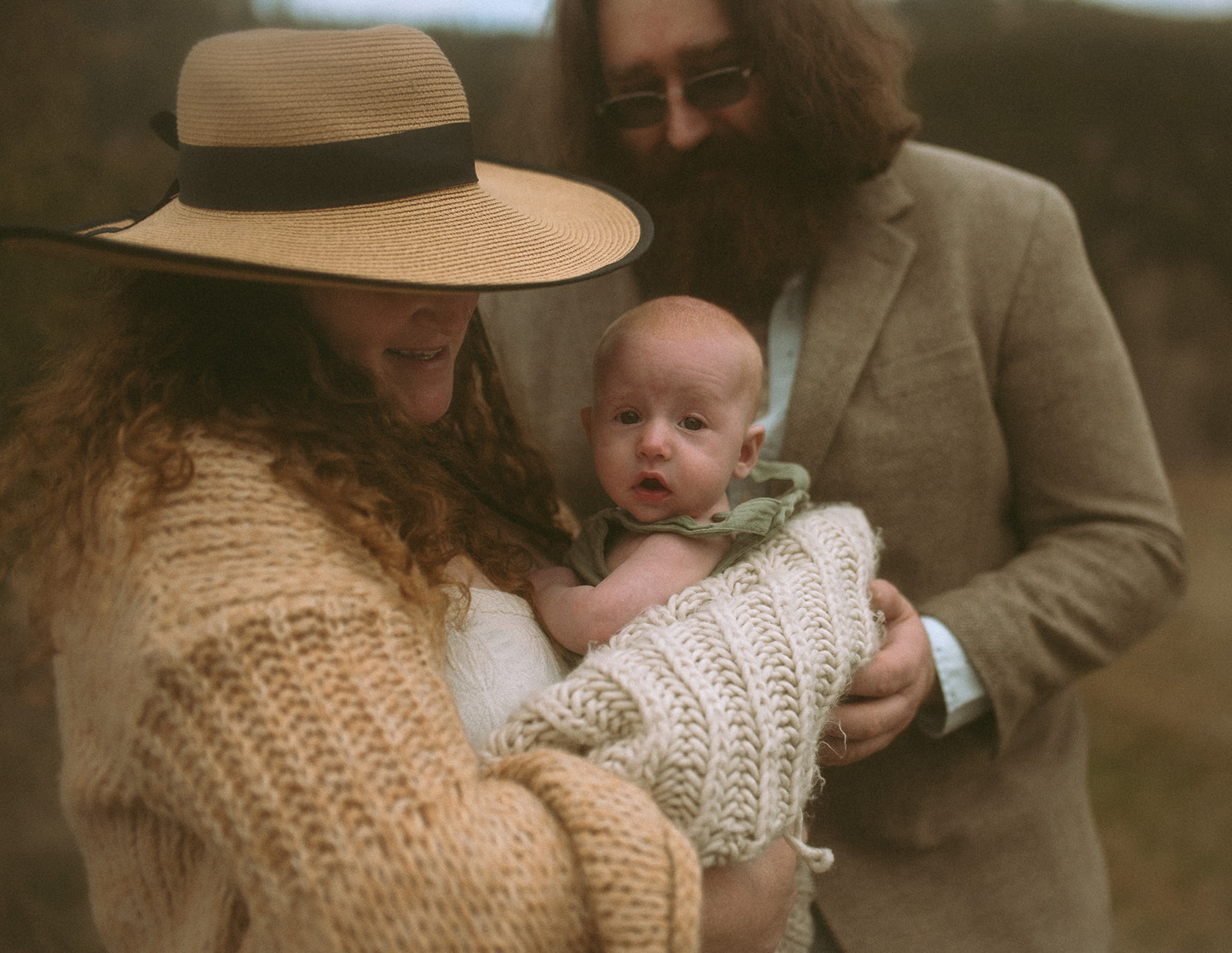 Happy mom and dad snuggle their infant in a knit blanket while walking outdoors in another knit sweater, wide brim hat and tweed jacket