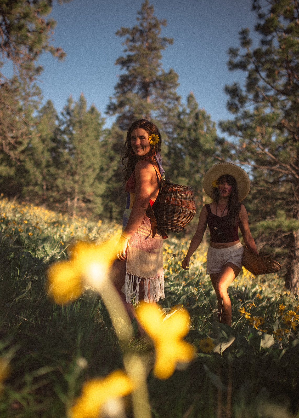 Two women walk with baskets through a field of flowers with one in their hair
