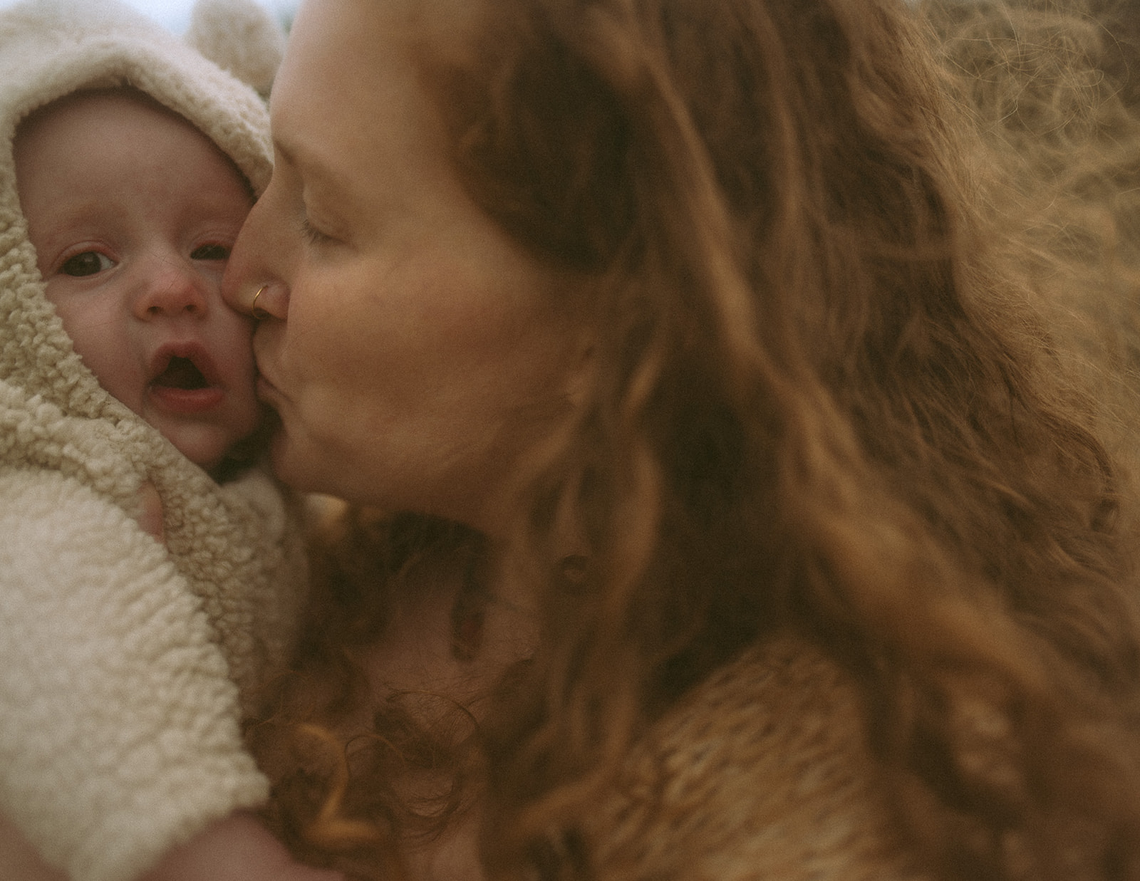 A happy mother with red hair kisses the cheek of her infant in a wool sweater and hood after some pelvic floor therapy in Hamilton, MT