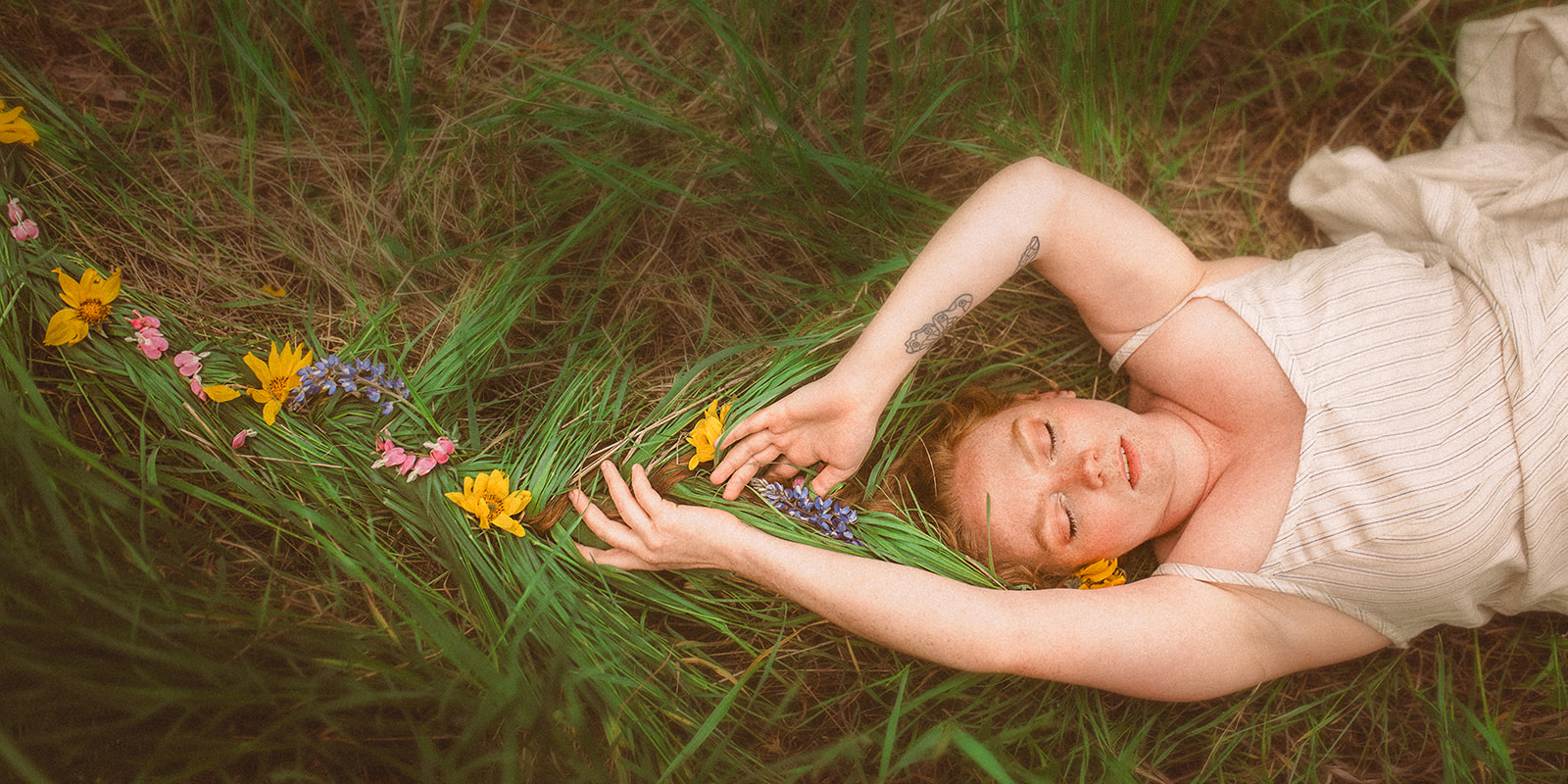 A pregnant woman with her hair braided into the long grass lays in a field in a white maternity dress