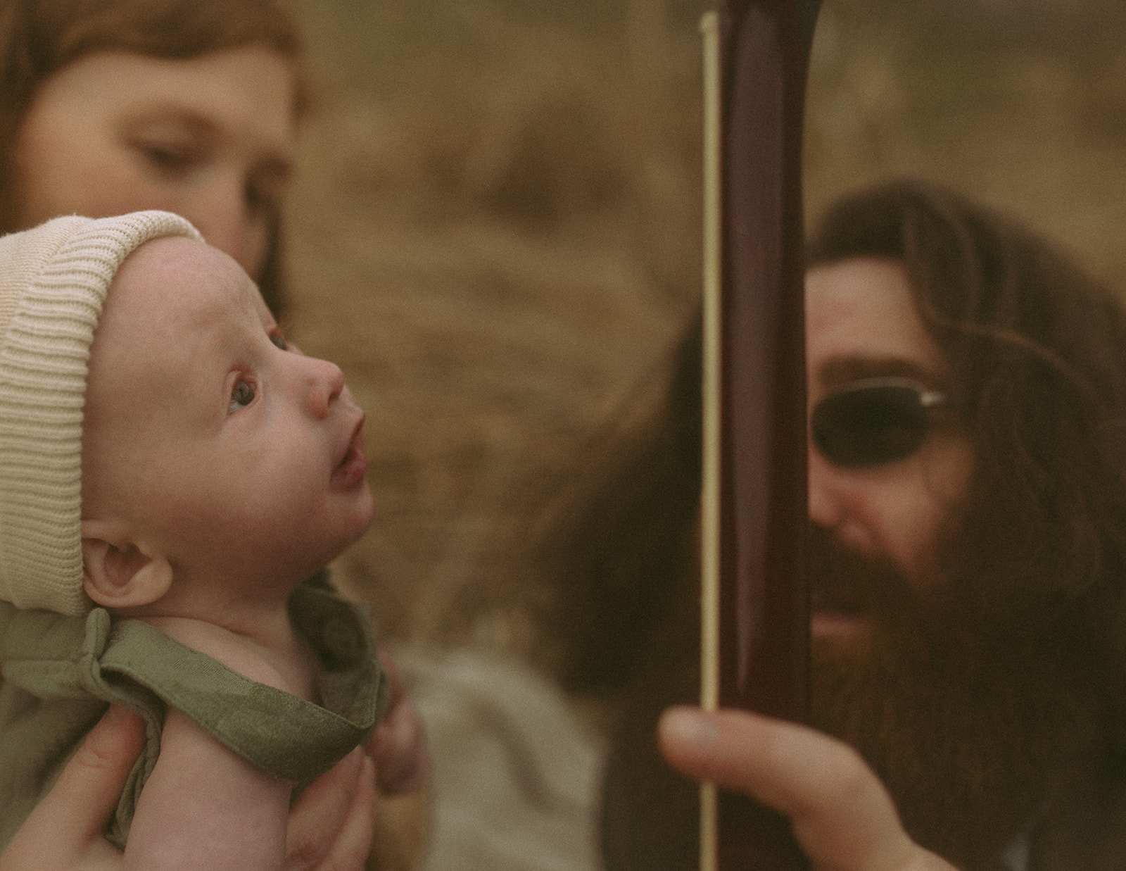 A baby examins a guitar neck held by dad in a field