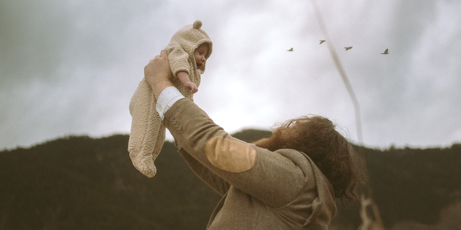 A happy new dad in a tweed jacket lifts his baby in a teddy onesie in a field as birds fly over after visiting playgrounds in Hamilton, MT