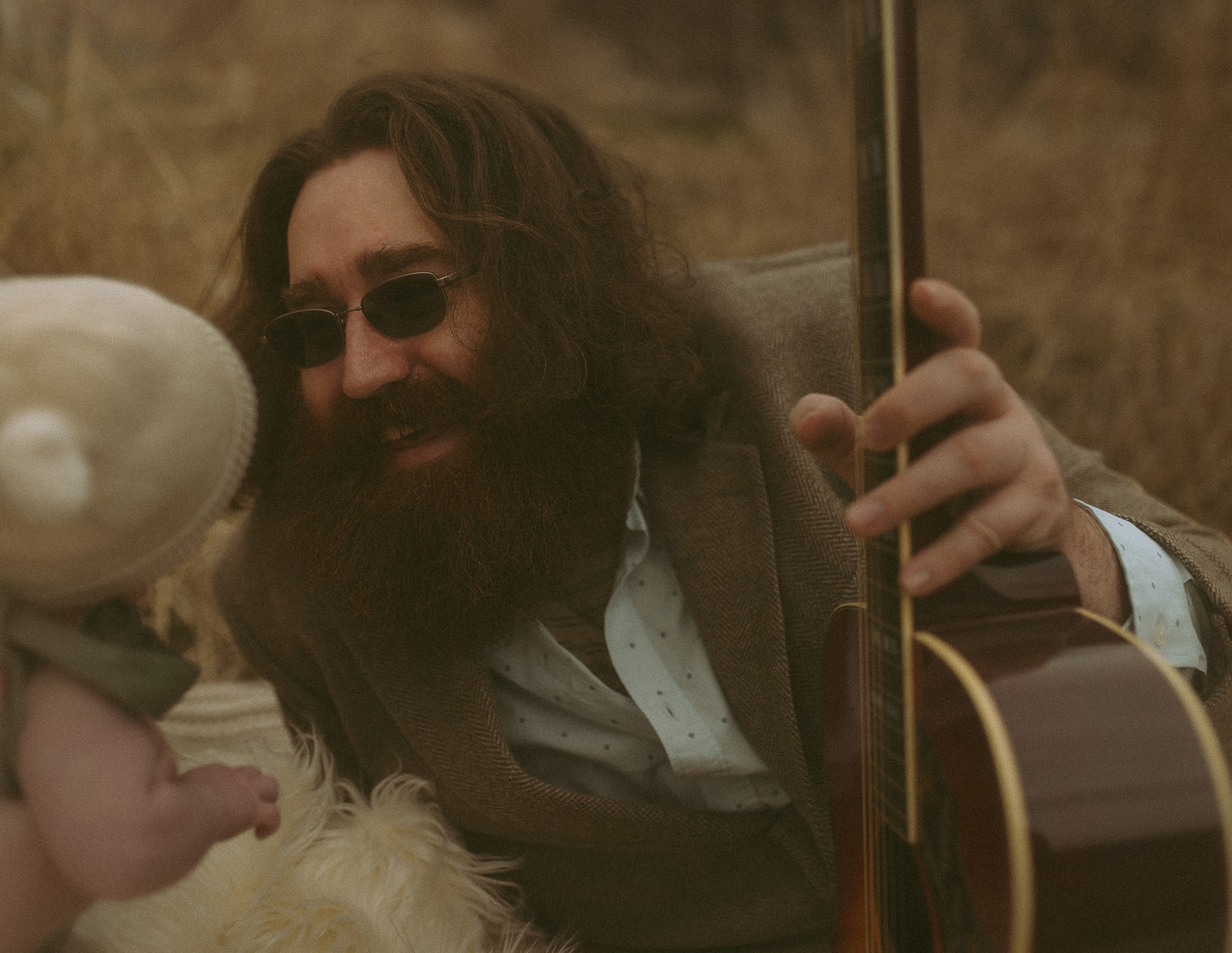 A happy new dad with a big beard and guitar smiles at his baby while sitting in a field after visiting playgrounds in Hamilton, MT