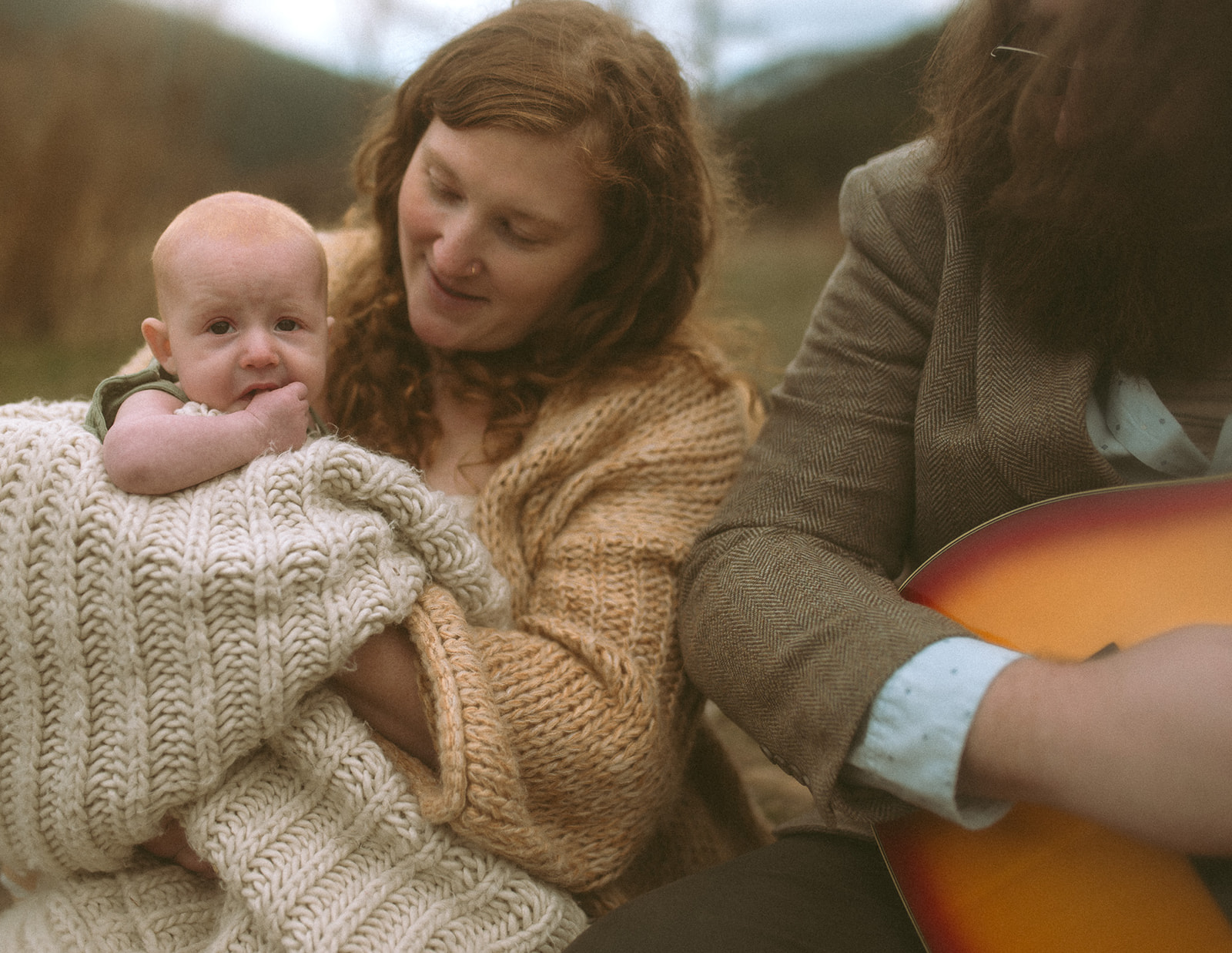 A mom smiles down at her infant in her lap wrapped in a white knit blanket while sitting in a meadow next to dad playing a sunburst guitar in a suit
