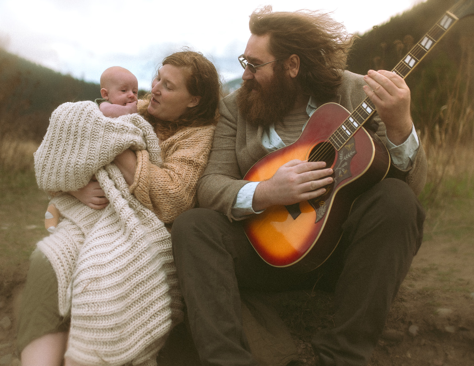 A happy mom holds her infant in a large blanket while sitting with guitar playing dad thanks to a prenatal chiropractor in Hamilton, MT