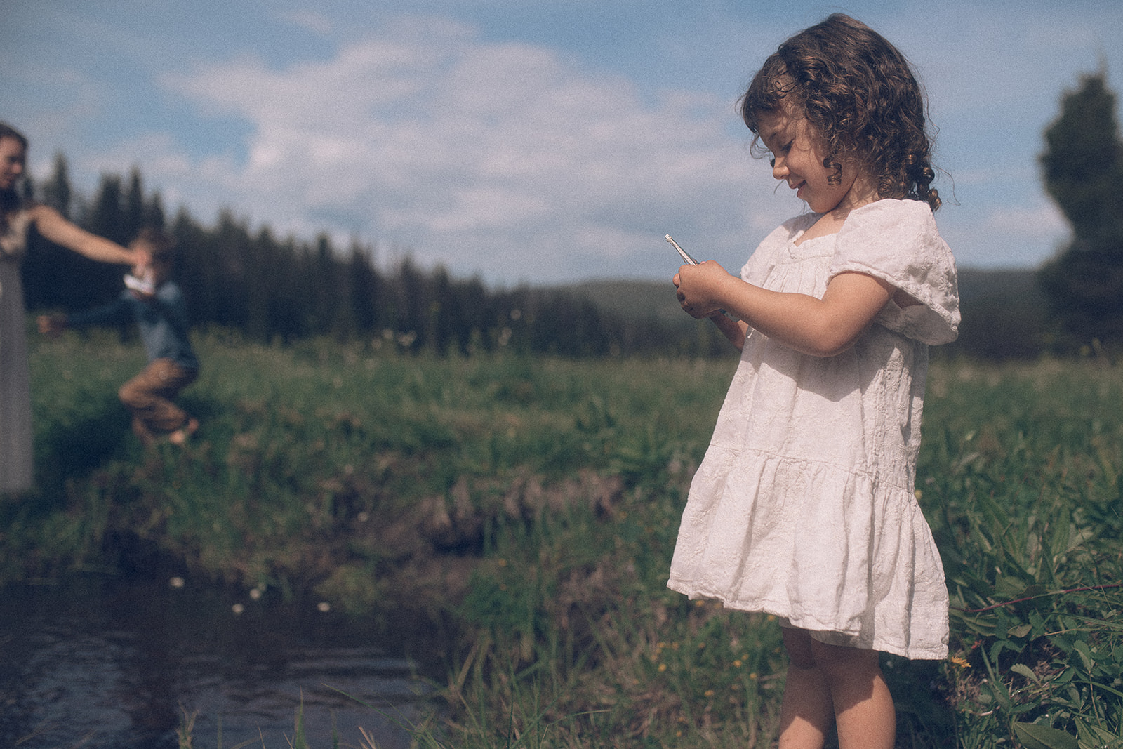 A toddler girl exams a tiny toy boat while standing on the edge of a creek in a meadow in a white dress