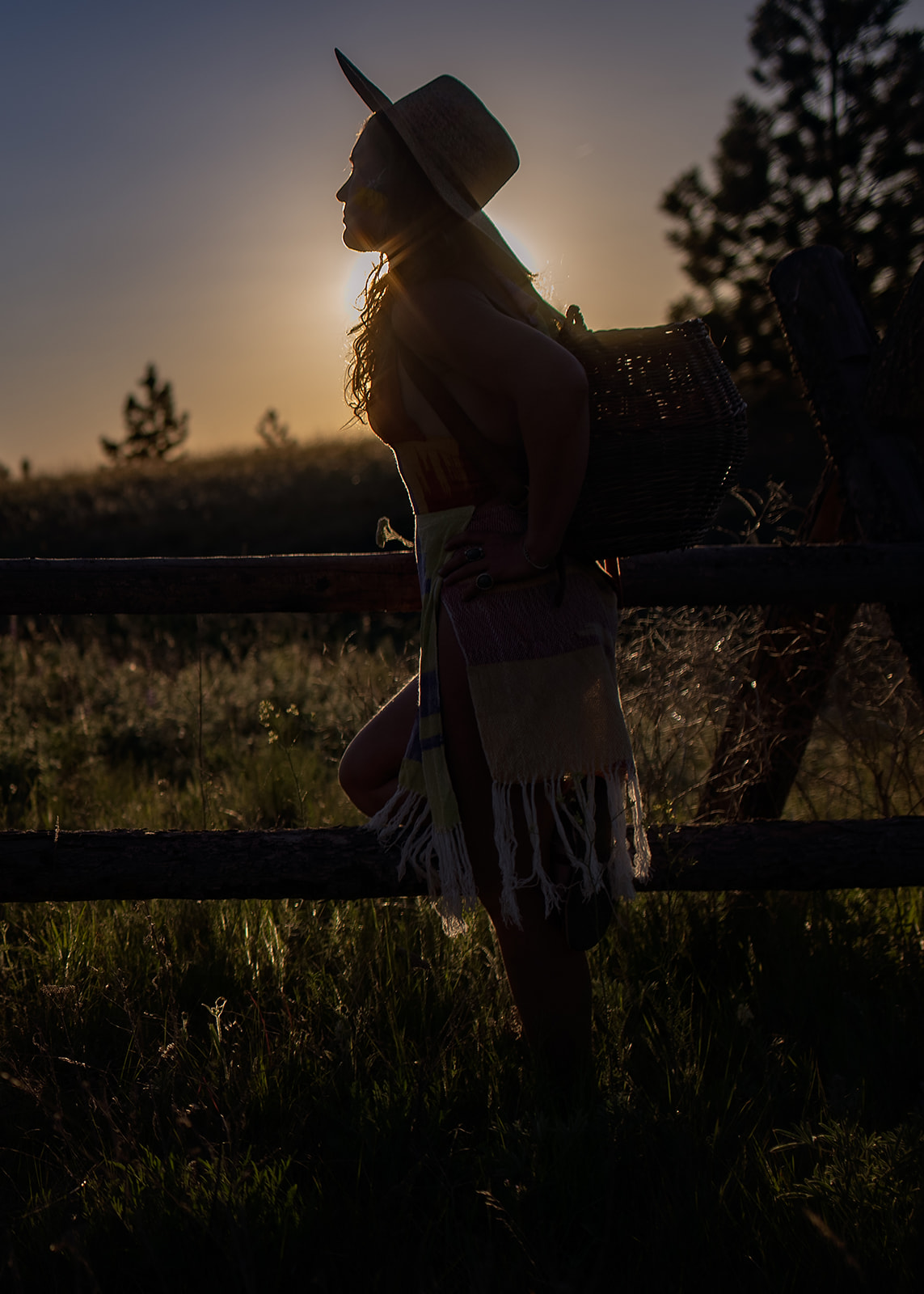 Silhouette of a woman in a big hat leaning on a pasture fence at sunset