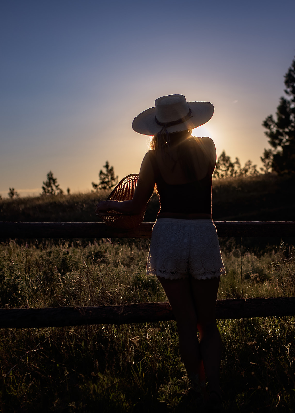 A woman leans on a pasture fence in a big brim hat watching the sunset after attending a private school in Hamilton, MT