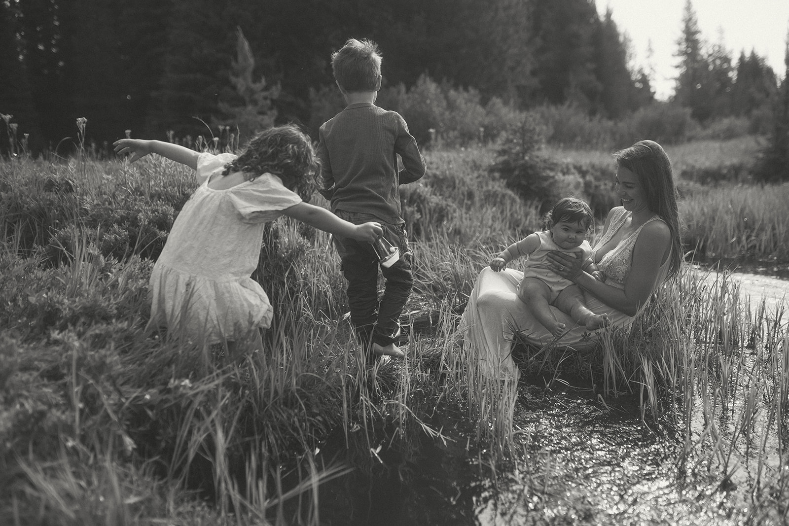 A happy mom sits on the edge of a small creek with a baby in her lap as her two toddlers play around them