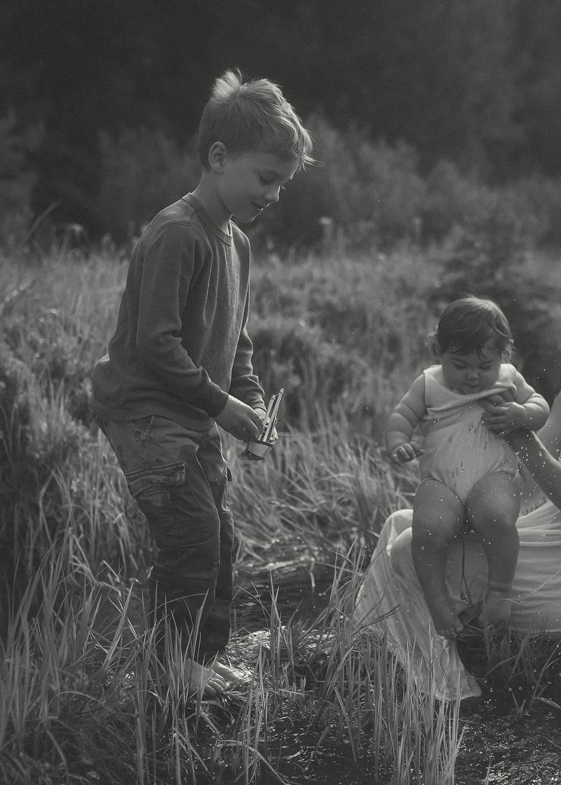 A young boy plays with a toy boat in a shallow creek with mom and baby sibling after visiting splash pads in Hamilton, MT