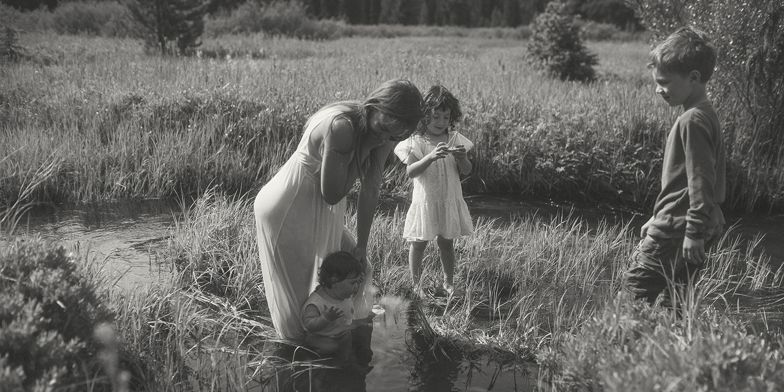 A mother in a white dress plays with her three young kids in a shallow creek before visiting splash pads in Hamilton, MT
