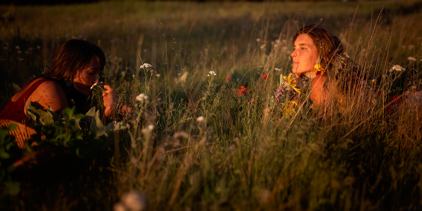 Two women lay in a field of colorful wildflowers at sunset smelling them