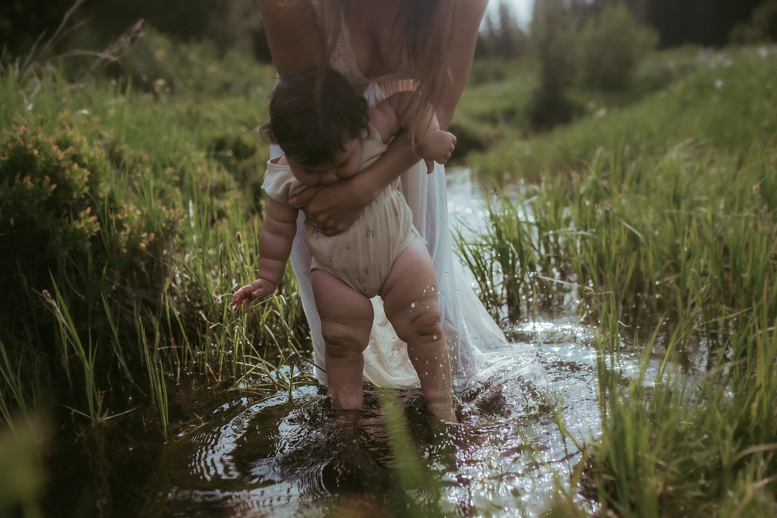 A mom helps her baby play in a shallow grassy creek in a white onesie and white dress