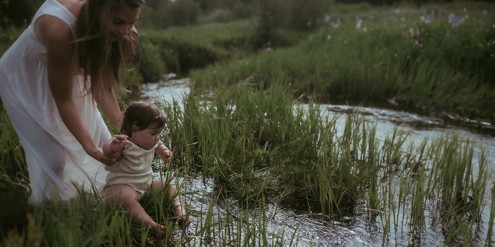 A mom sits her baby on the edge of a shallow creek in a white dress after some swim lessons in Hamilton, MT