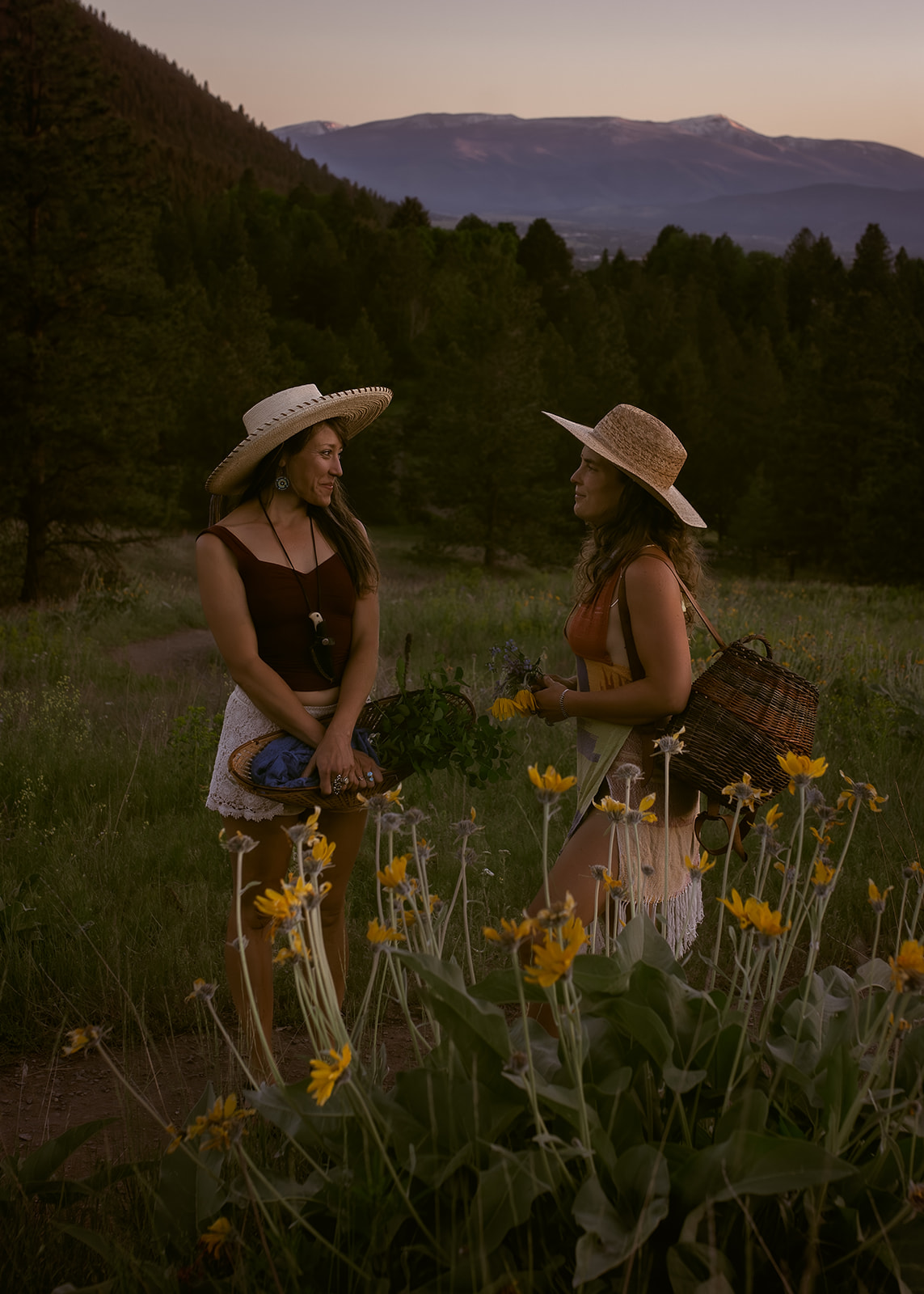 Two women chat on a trail by some wildflowers at sunset with large woven hats