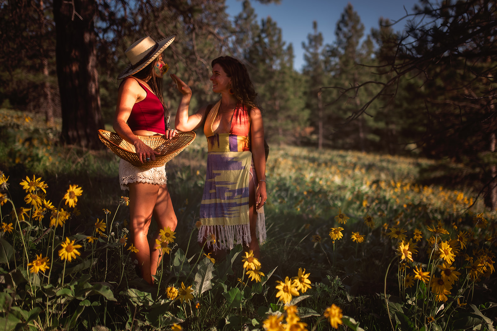 Two women in light clothing laugh and play in a field of yellow wildflowers at sunset with a woven basket before visiting pumpkin patch in Hamilton, MT