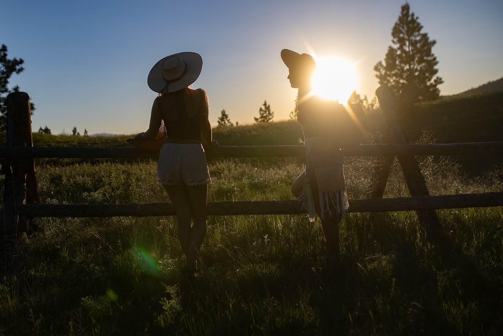 Silhouette of two women in shorts and large brimmed hats leaning on a wooden fence at sunset