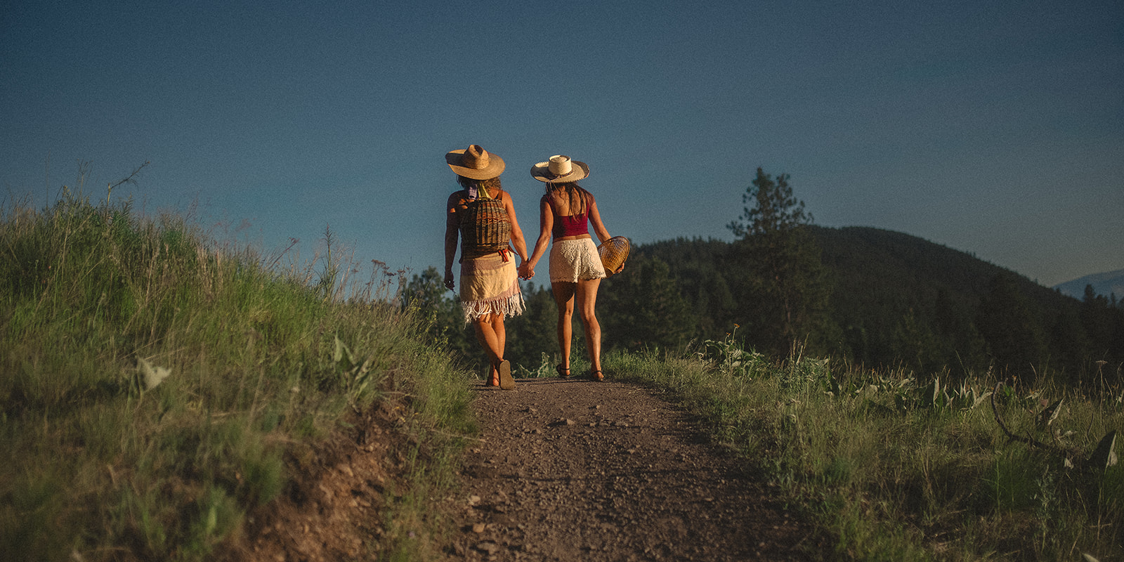 two women hold hands while hiking a trail at sunset in large hats after visiting a pumpkin patch in Hamilton, MT
