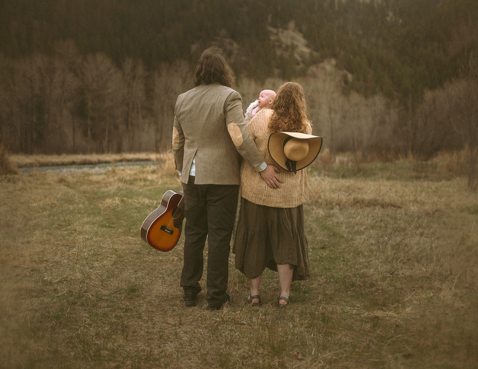 New mom and dad stand holding a guitar and their newborn while watching a sunset in the mountains before visiting Missoula Fall Events