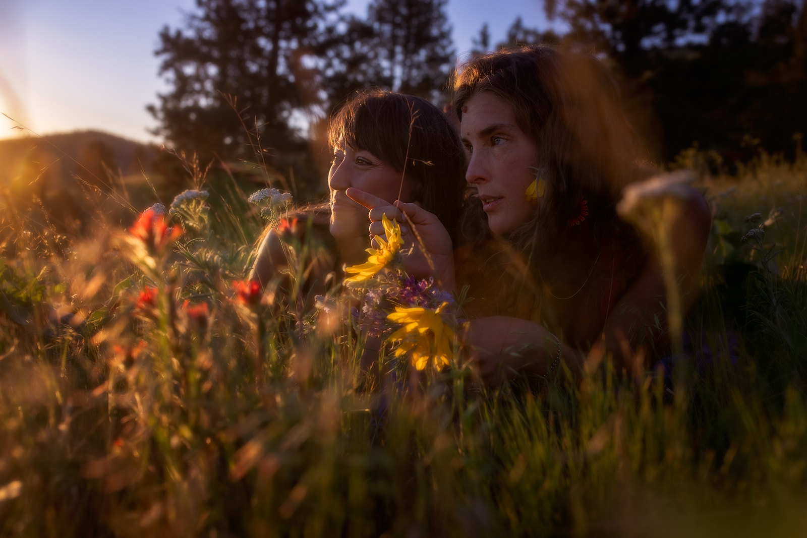 A couple cuddle among colorful wildflowers in the grass at sunset pointing to the sunset