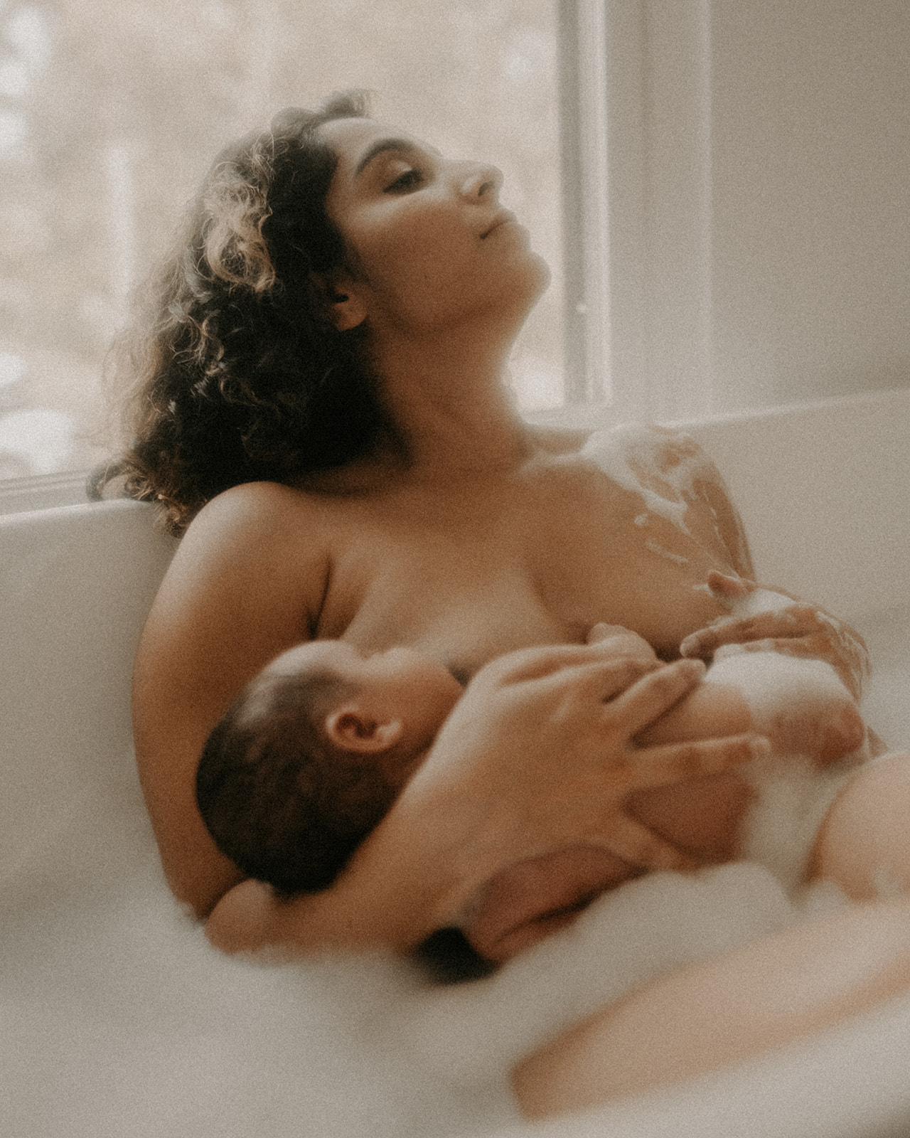 A mother basks in the sun of a window in a bubble bath breastfeeding her newborn thanks to Roots Birth Center Missoula