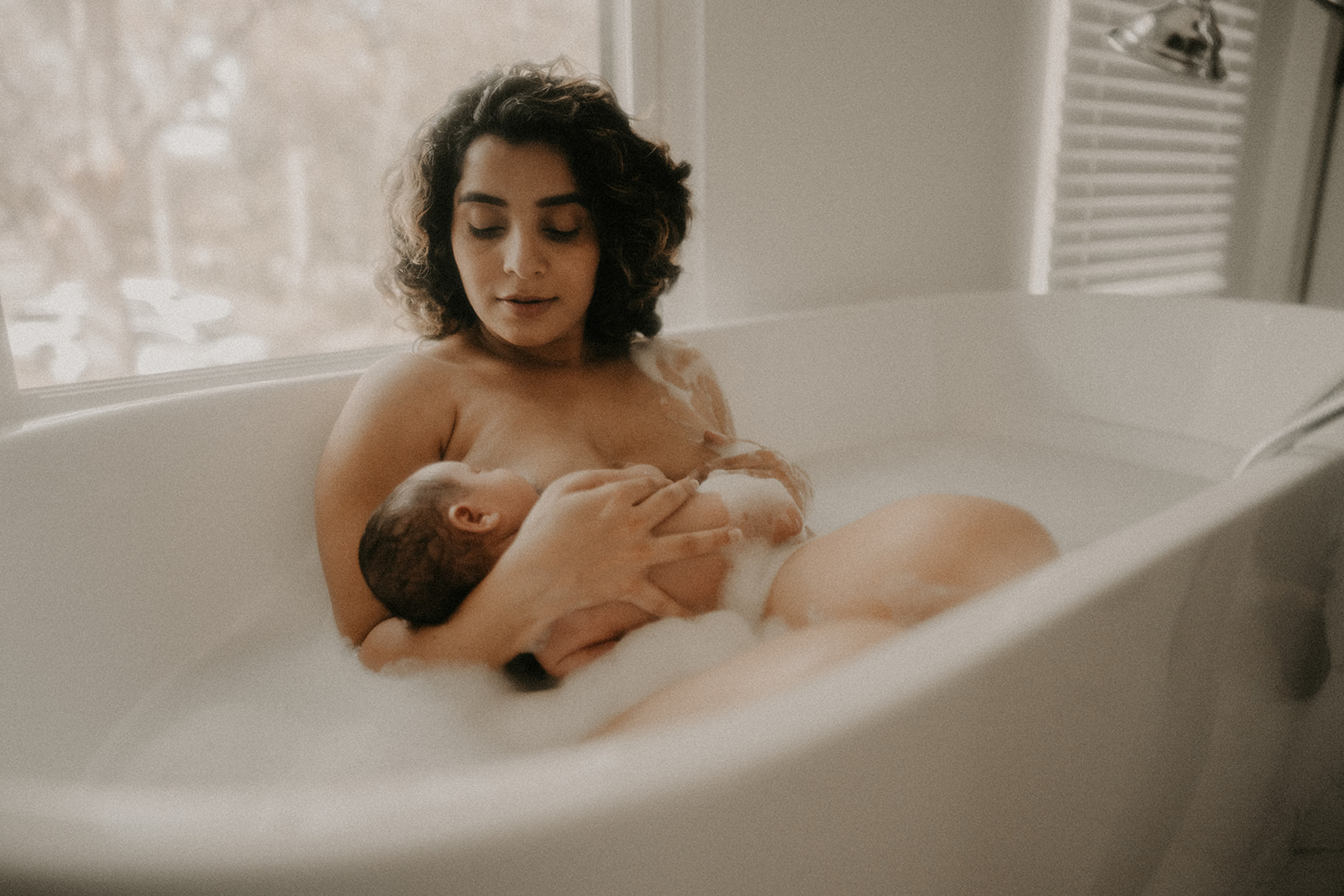 A new mother sits in a bubble bath under a window cradling her sleeping newborn on her chest thanks to Roots Birth Center Missoula