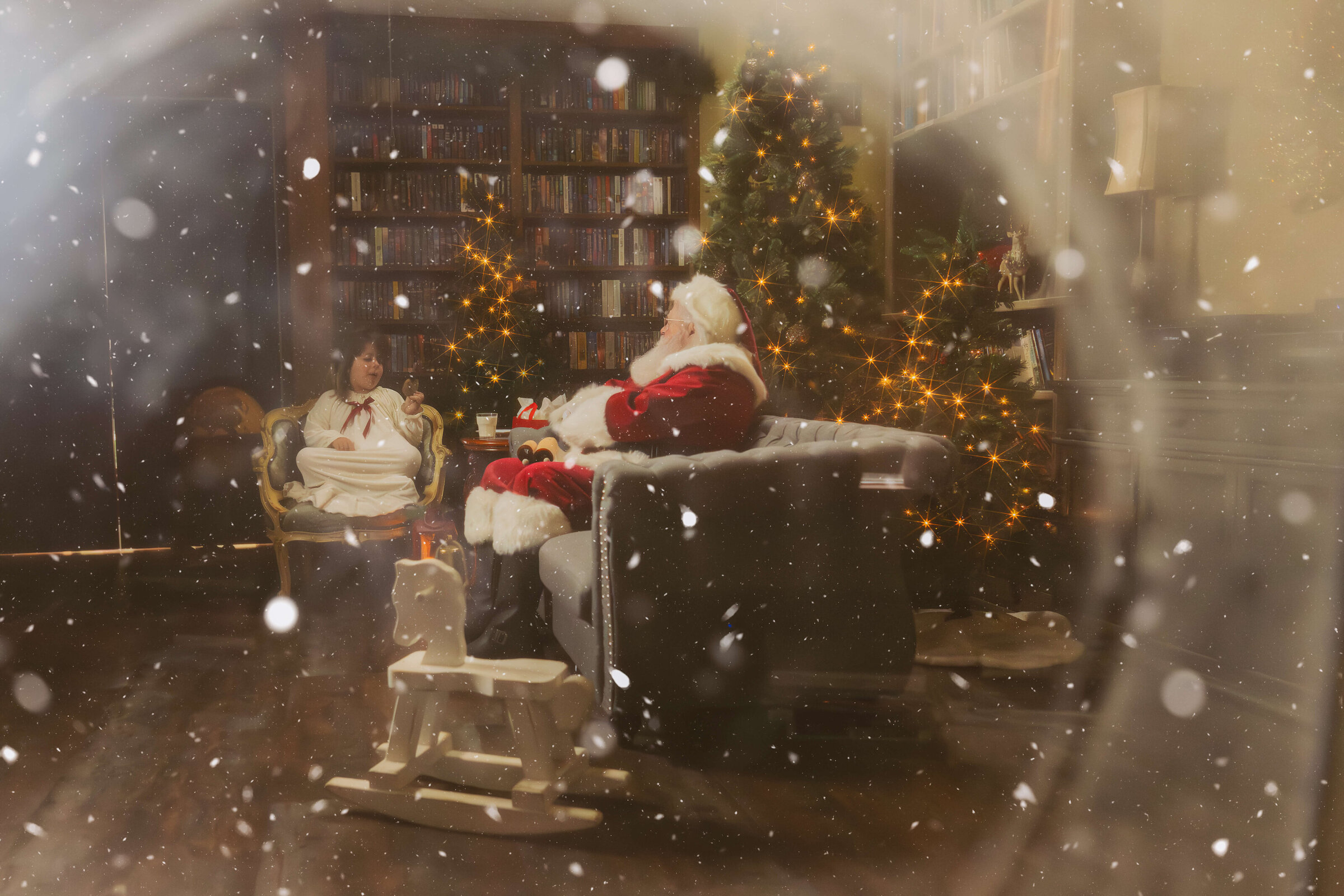 A young girl in white dress sits eating cookies in a living room with santa in a magical snowy scene after Christmas events in Hamilton, MT