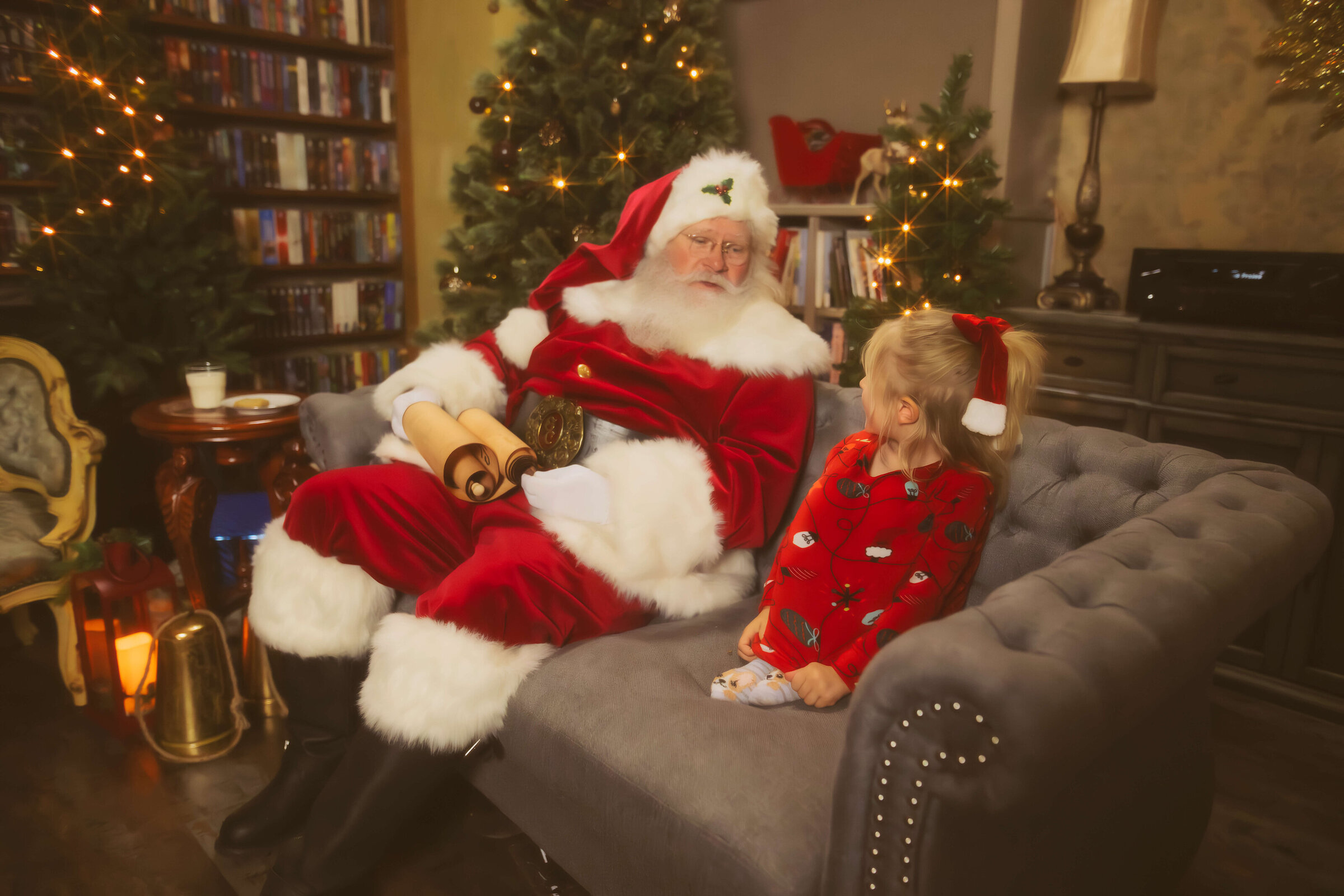 A Santa sits on a grey couch chatting with a toddler girl in red christmas pajamas while holding a scroll