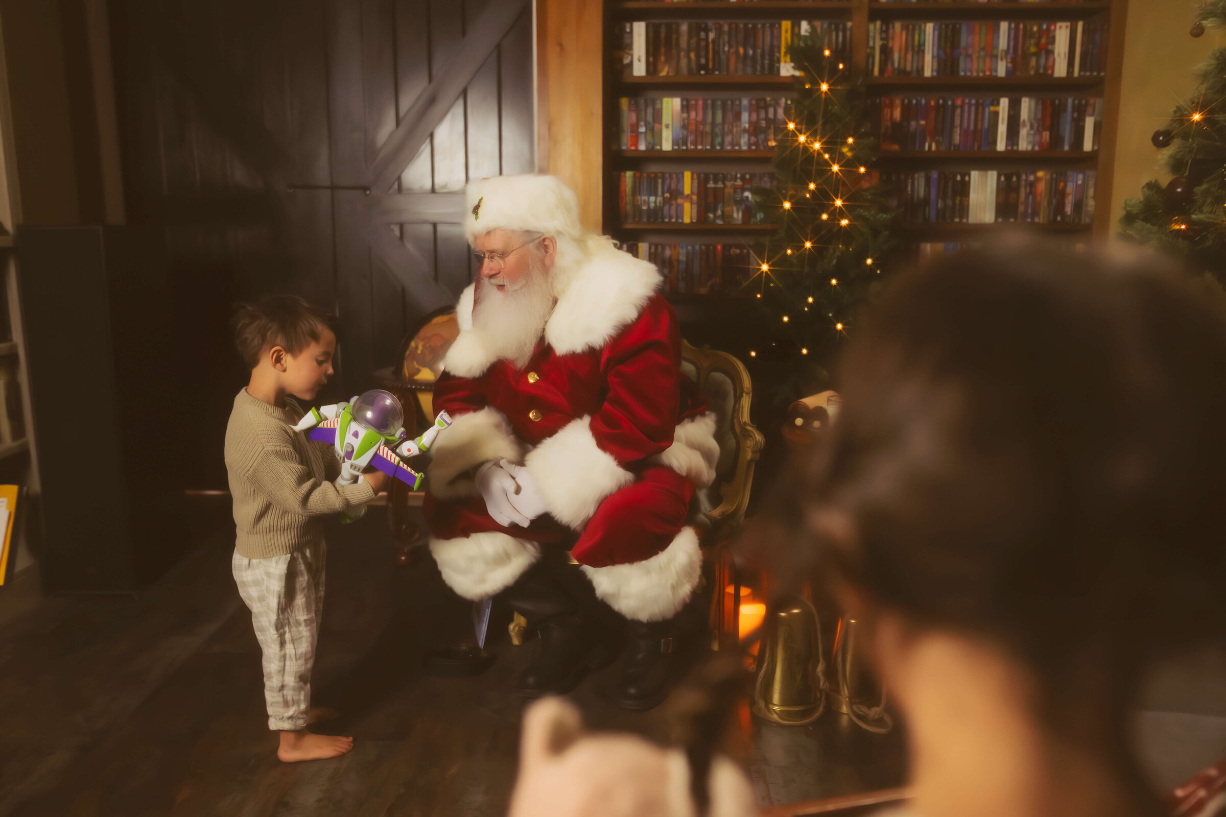 A toddler boy in a sweater and plaid pants shows Santa one of his toys in a living room