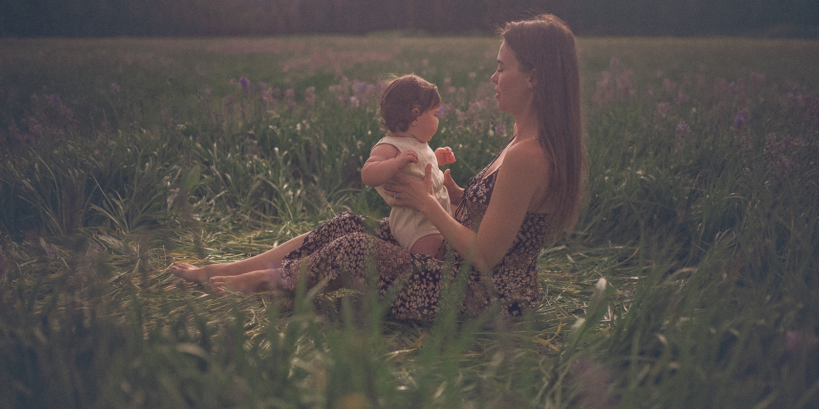A mother in a floral print dress plays in the wildflowers with her baby in her lap at sunset