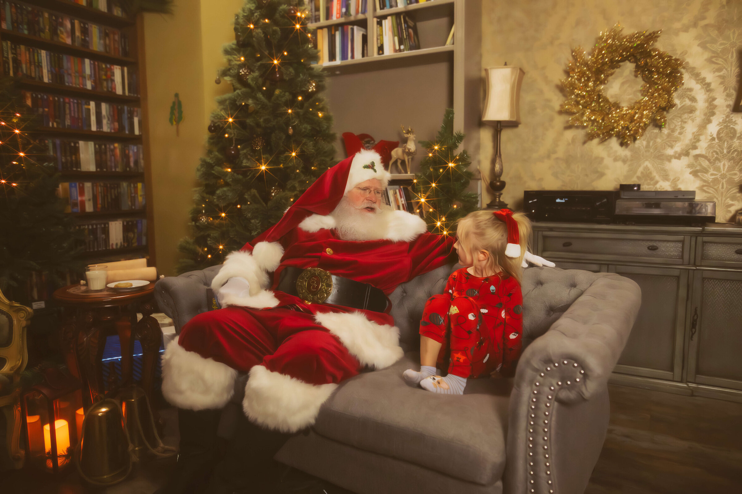 a santa in hamilton, mt sits on a grey couch in a living room talking to a young girl in red pajamas