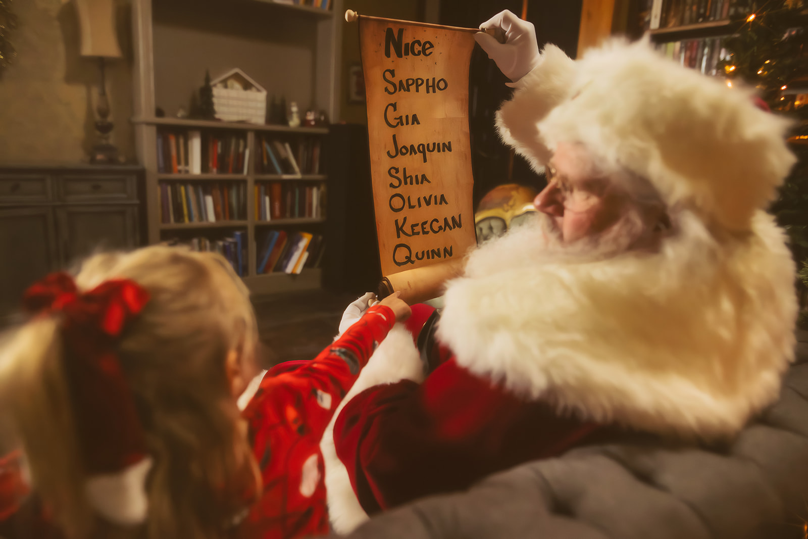 A Santa in Missoula, MT reads his nice list to a toddler girl in holiday pajamas while sitting on a couch