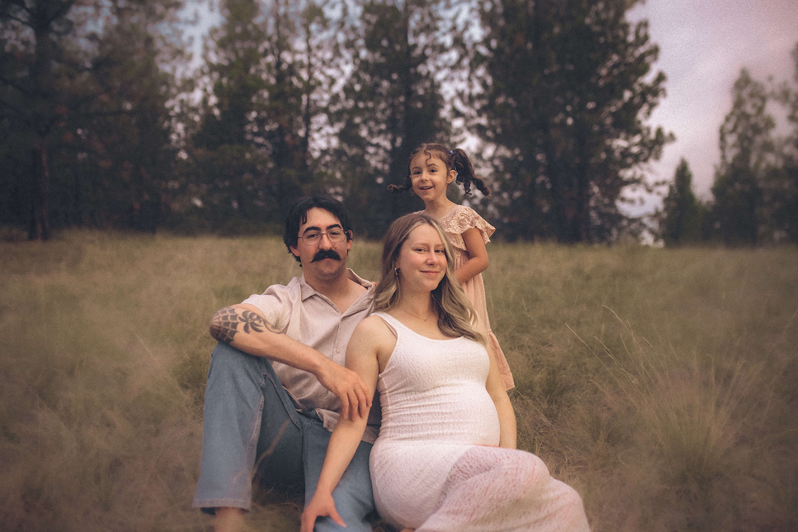 Happy expecting mom and dad sit in the grass in a meadow with their toddler daughter playing around them at sunset