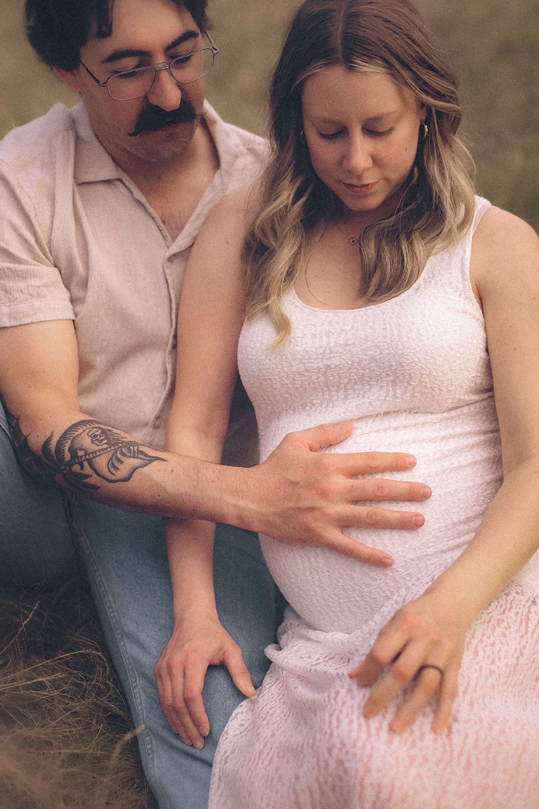 Parents to be snuggle in the grass with hands on the bump in white shirt and dress after meeting Fern Creek Midwives