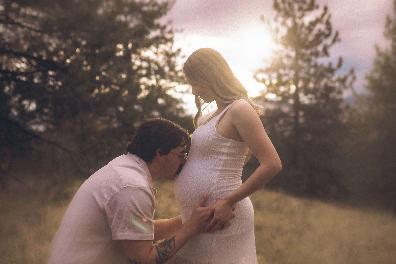 A man in a white shirt kisses the pregnant belly of his wide while in a forest at sunset after meeting Fern Creek Midwives