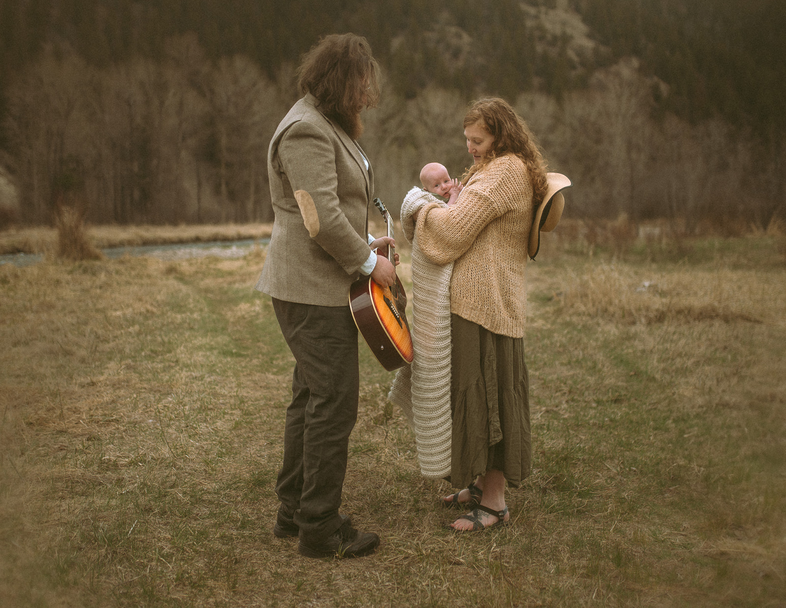 A mom cradles her awake newborn as dad holds an orange guitar on a meadow by a river