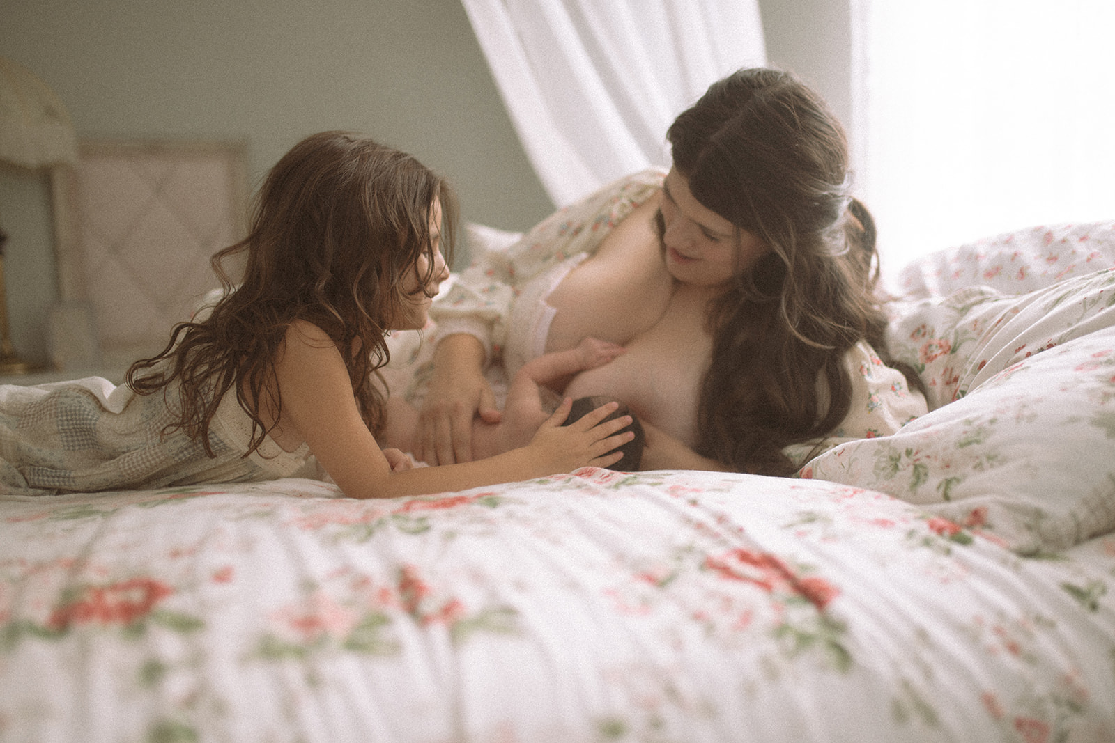 a mother in a floral print dress breastfeeds her newborn while laying across a bed as her toddler daughter rests her hand on the newborn's head thanks to a lactation consultant in Polson, MT