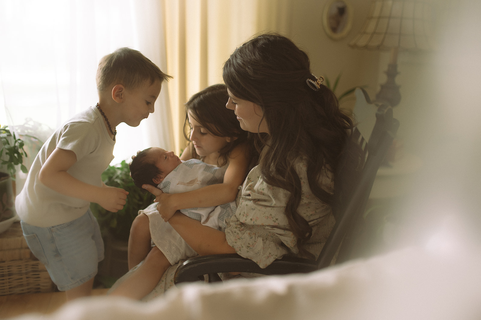 A mother helps her toddler daughter hold her newborn in her lap in a wooden rocking chair as big brother looks on after visiting pediatricians in Polson, MT