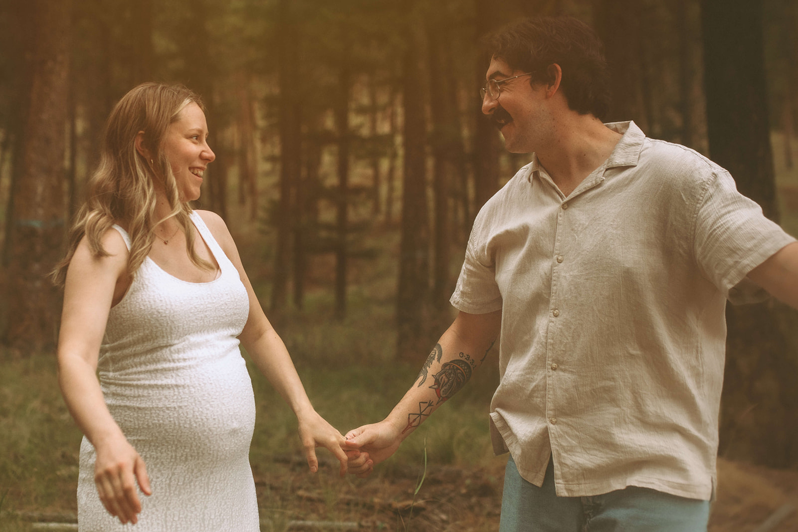 A happt pregnant couple play in a forest in white outfits