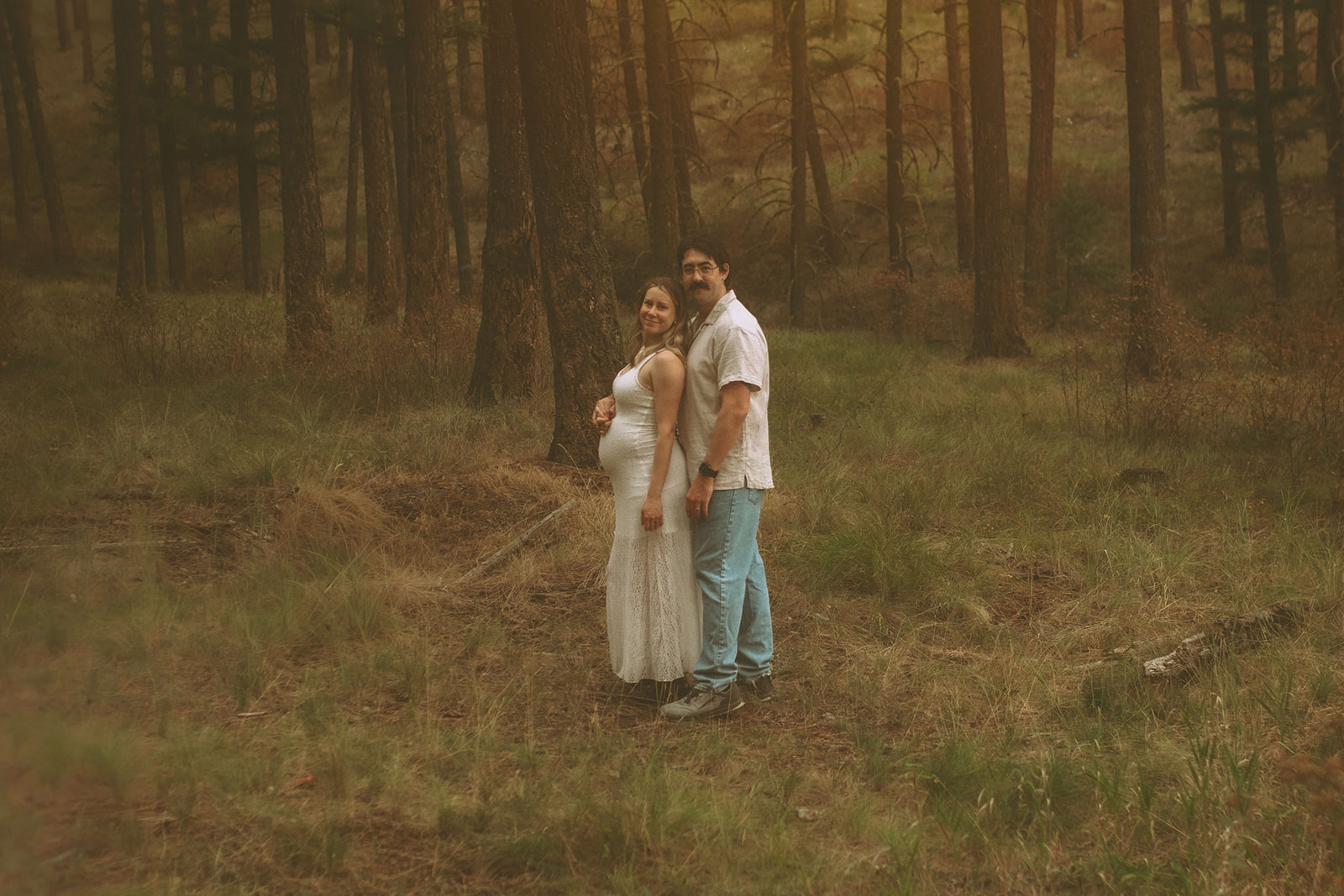 A smiling expecting couple stand in a forest at sunset in white wit hands on the bump after visiting baby shower venues in Polson, MT